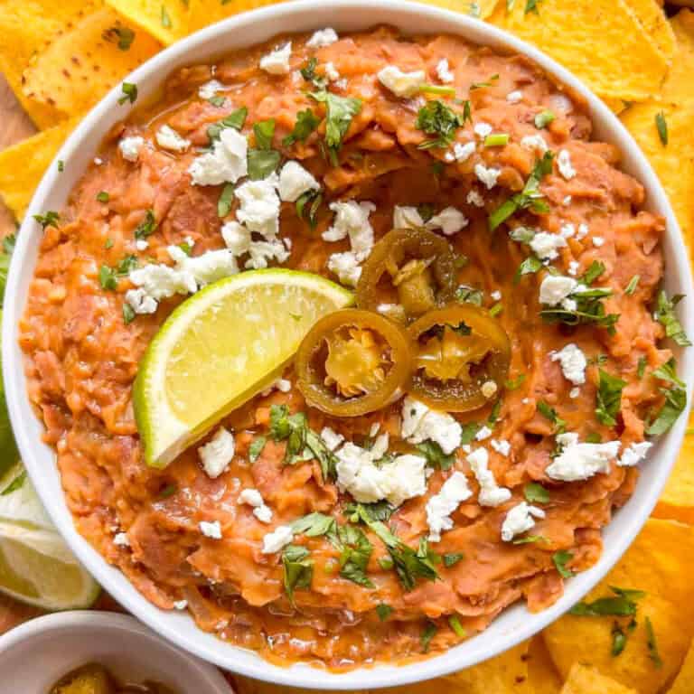 a bowl of refried beans surrounded by toritlla chips and lime wedges. The refried beans are topped with crumbled feta, jalapenos, a lime wedge, and fresh chopped cilantro