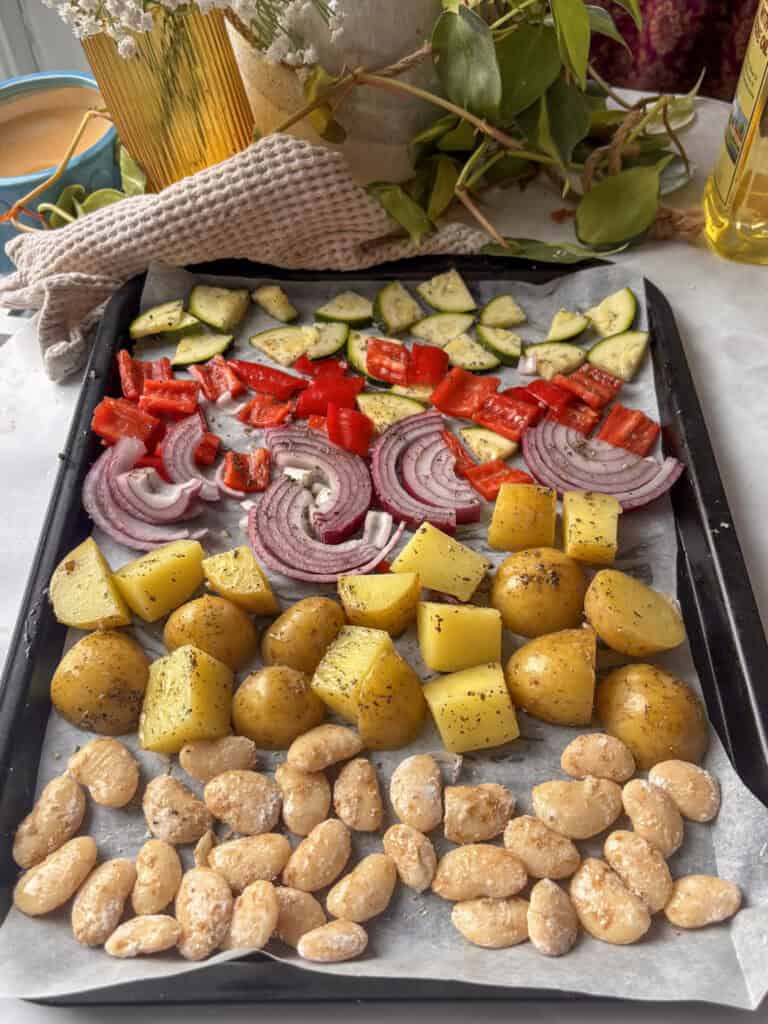 a baking tray lined with zucchini, red pepper, red onion, potatoes, and butter beans tossed in Italian seasonings ready to be roasted.