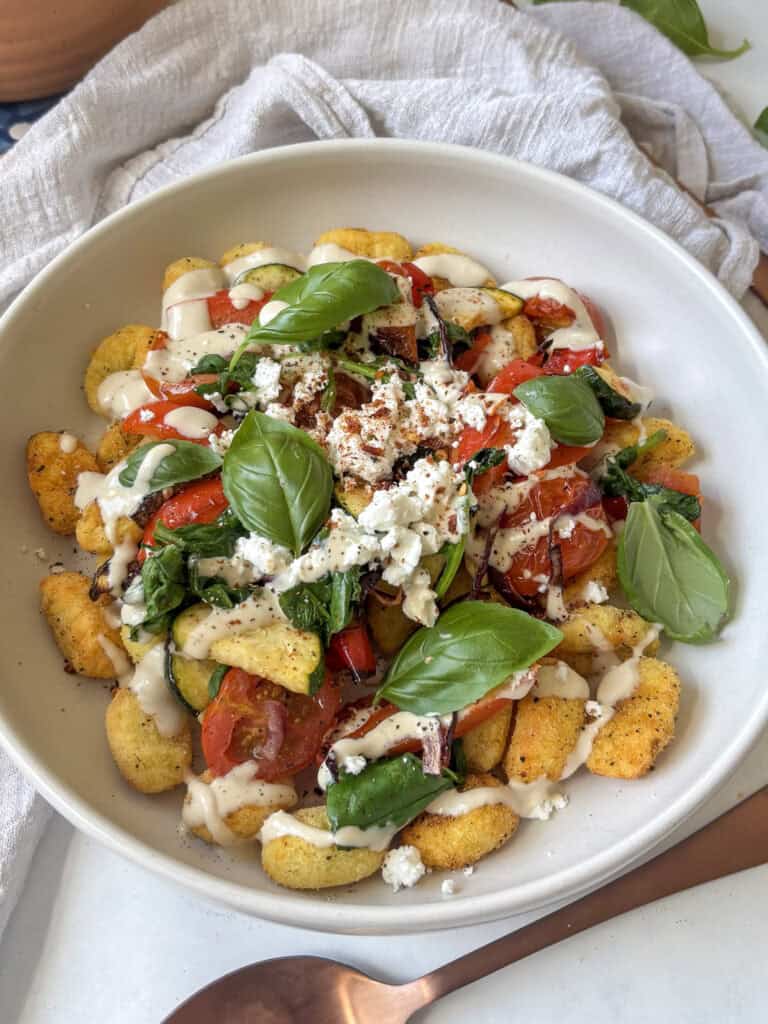 a bowl of baked gnocchi with roasted vegetables, feta, and a creamy tahini sauce, topped with fresh basil leaves