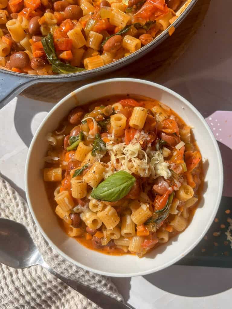 a white bowl of pasta e fagioli topped with cheese and basil