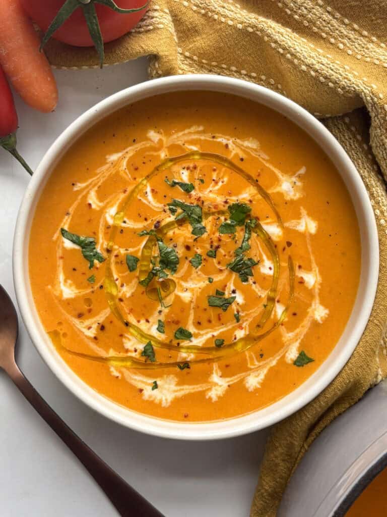 a bowl of butternut squash and red pepper soup topped with cream, olive oil, and fresh herbs and seasoning