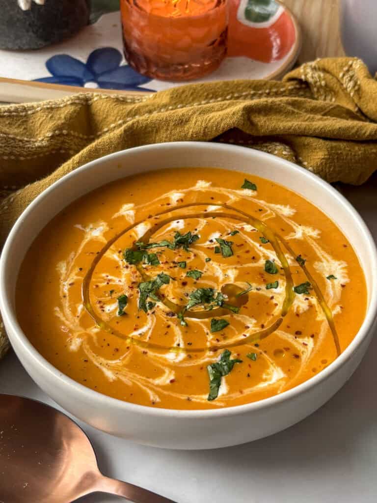 a bowl of butternut squash and red pepper soup topped with cream, olive oil, and fresh herbs and seasoning