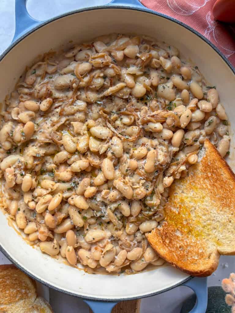 a casserole dish of caramelized onions and white beans topped with rosemary, with a slice of toasted sourdough