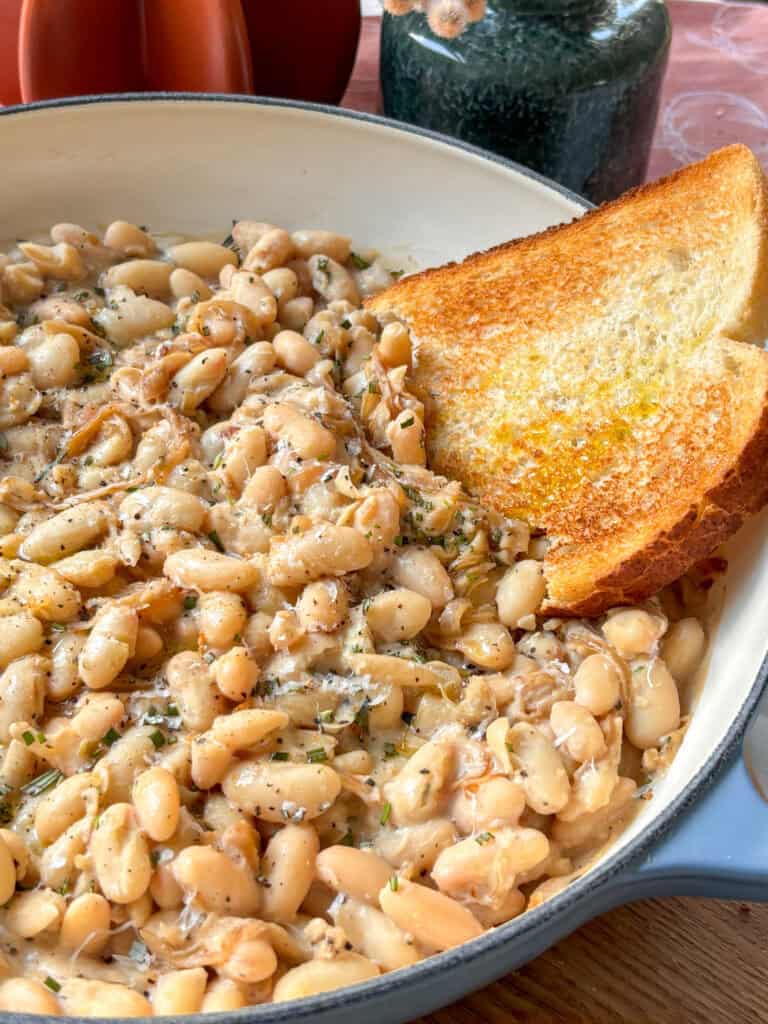 a casserole dish of caramelized onions and white beans topped with rosemary and black pepper, with a slice of toasted sourdough