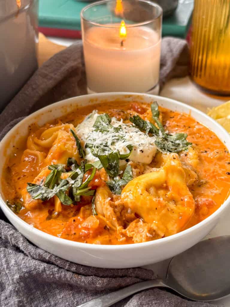 side on photo of creamy tortellini soup with cheese tortellini, white beans, crumbled sausage,in a creamy tomato sauce topped with ricotta and basil. A candle is in the background