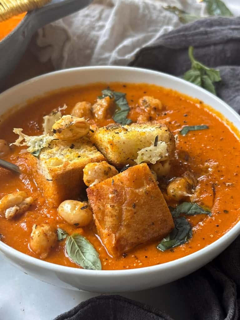 a side on photo of a bowl of spicy roasted red pepper soup topped with croutons, crunchy white beans, and basil