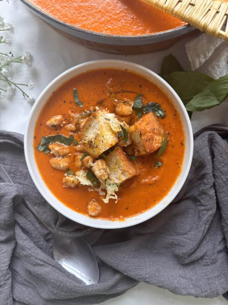 a bowl of spicy roasted red pepper soup topped with croutons, crunchy white beans, and basil