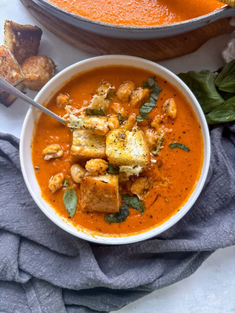 an overhead shot of a bowl of spicy roasted red pepper soup topped with croutons, crunchy white beans, and basil