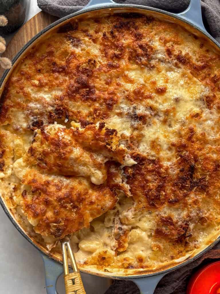 a photo of a dish of creamy baked mac and cheese, topped with a cheesy breadcrumb topping baked until crispy and golden. A serving spoon is taking a scoop out of the mac and cheese