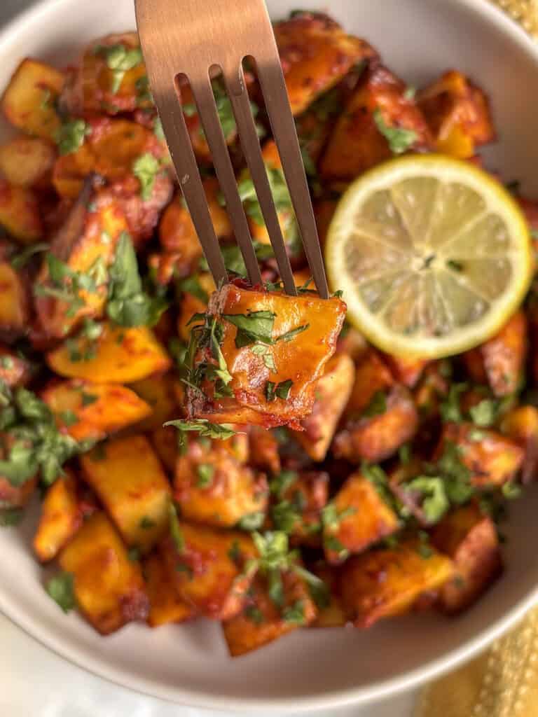 a fork spearing a cubed potato with herbs on it, with the bowl of batata harra in the bottom