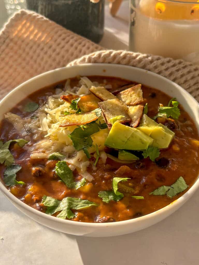 a close up of a bowl of creamy enchilada soup topped with avocao, cilantro, cheese, and trotilla chips