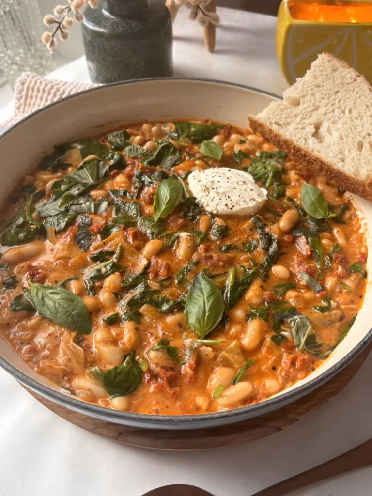 a skillet pan of creamy tuscan white beans with sun-dried tomatoes a, spinach, and basil with sour dough on the side
