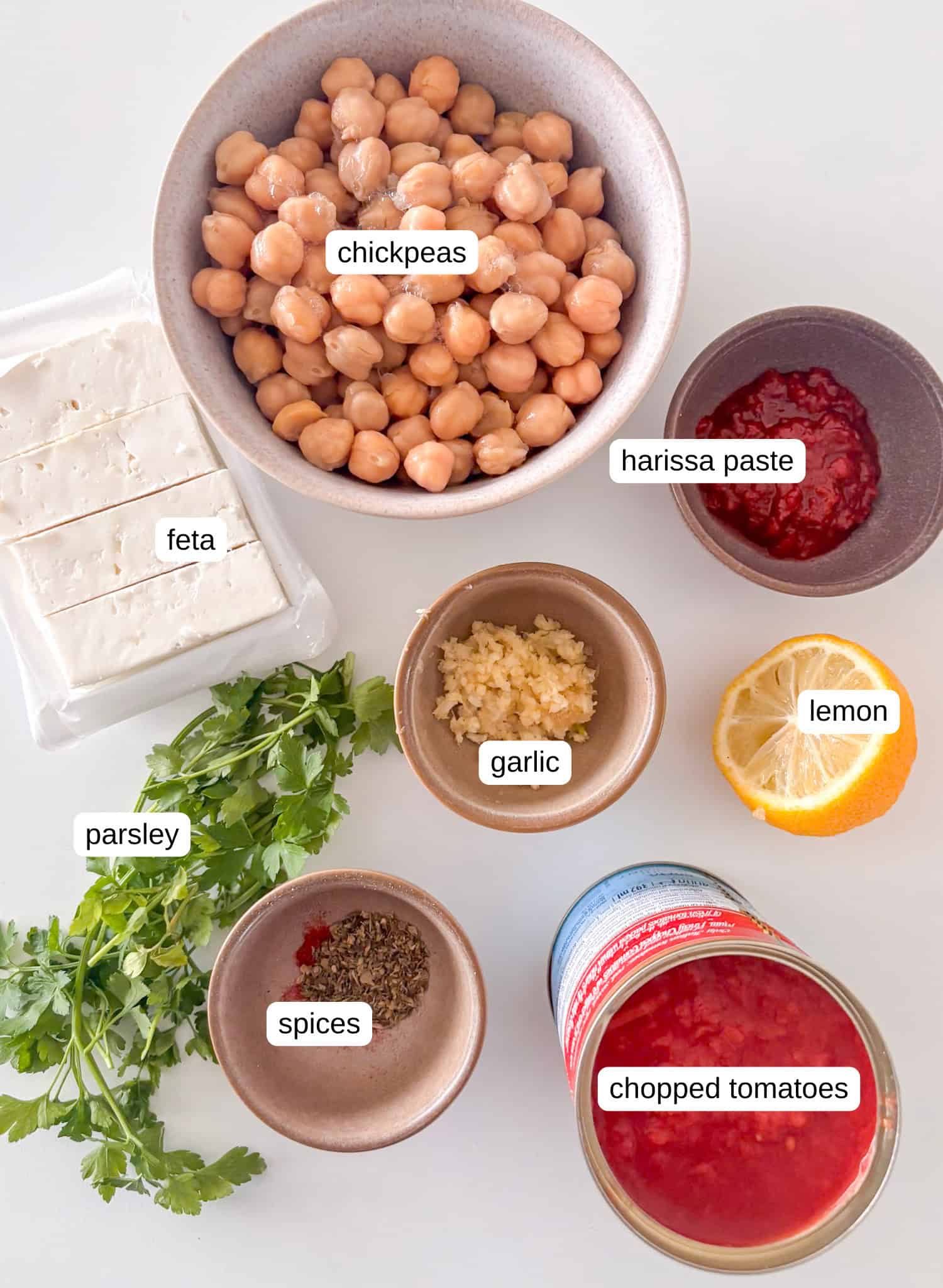 ingredients shot for these baked feta chickpeas: chickpeas, feta, harissa paste, garlic, lemon, parsley, spices, chopped tomatoes, lemon, olive oil