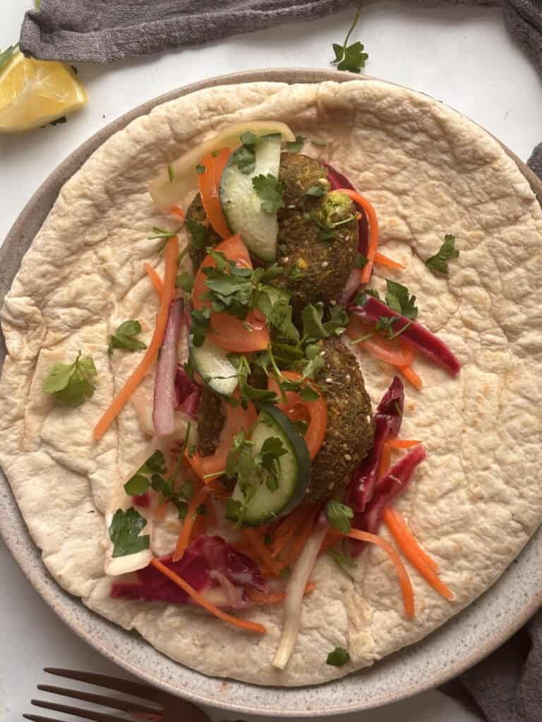 An open falafel wrap In the wrap, you can see falafels, cucumber, tomato, tahini sauce, and hot sauce