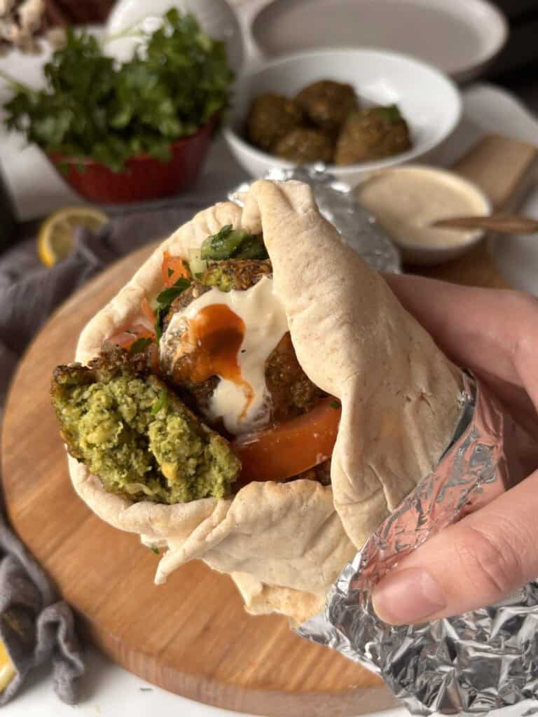 a falafel wrap on a wooden board, with foil holding them together. In the wrap, you can see falafels, cucumber, tomato, tahini sauce, and hot sauce