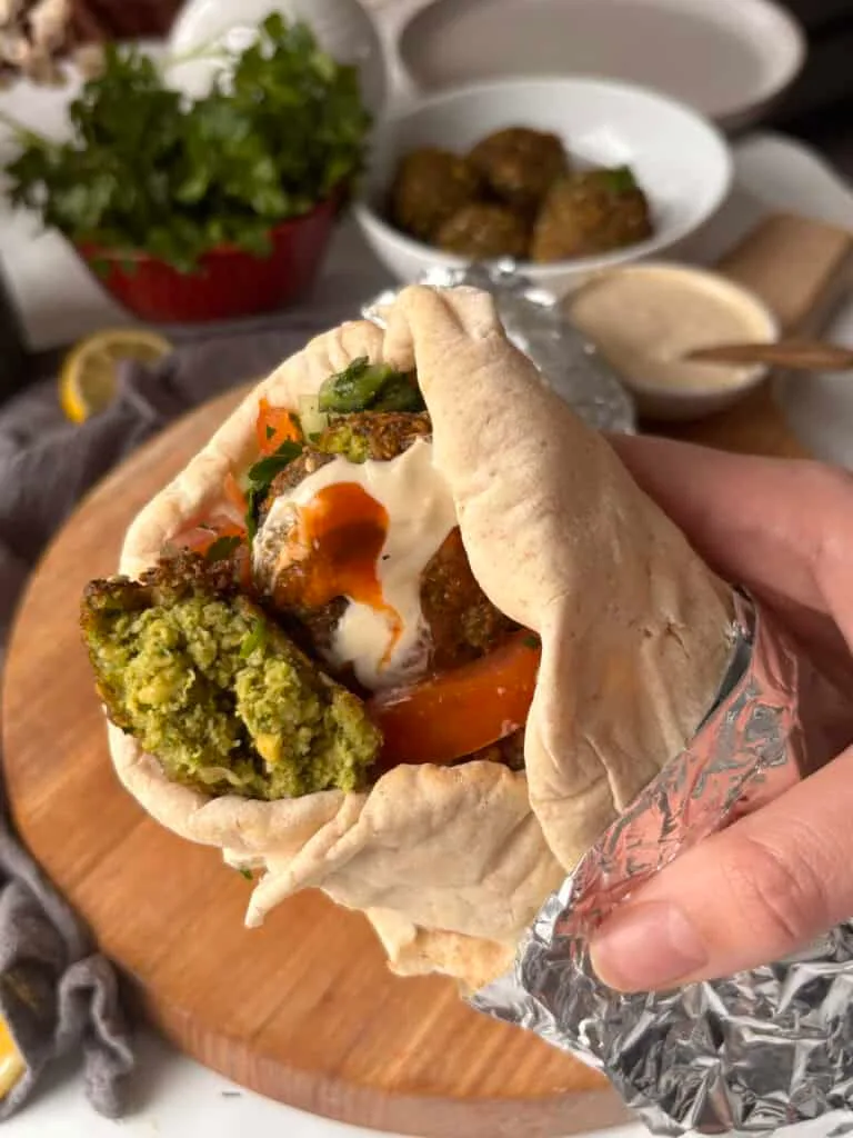 a falafel wrap on a wooden board, with foil holding them together. In the wrap, you can see falafels, cucumber, tomato, tahini sauce, and hot sauce