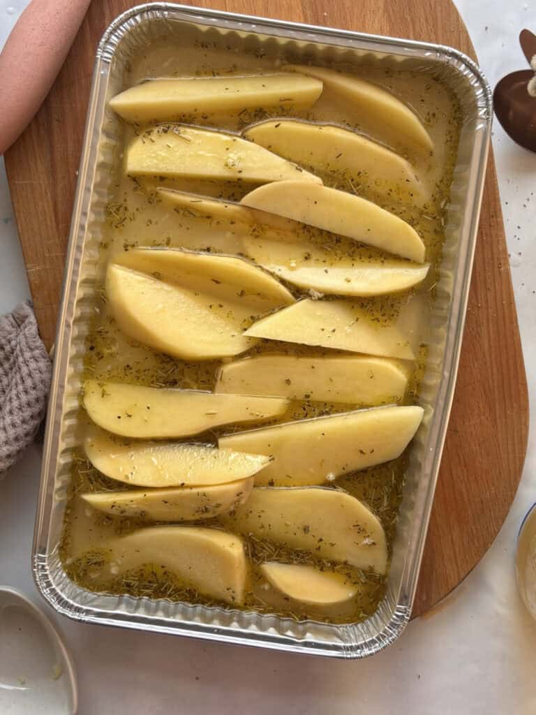 uncooked potatoes cut into wedge shapes in a foil roasting tin, with the lemony herby broth poured over them.