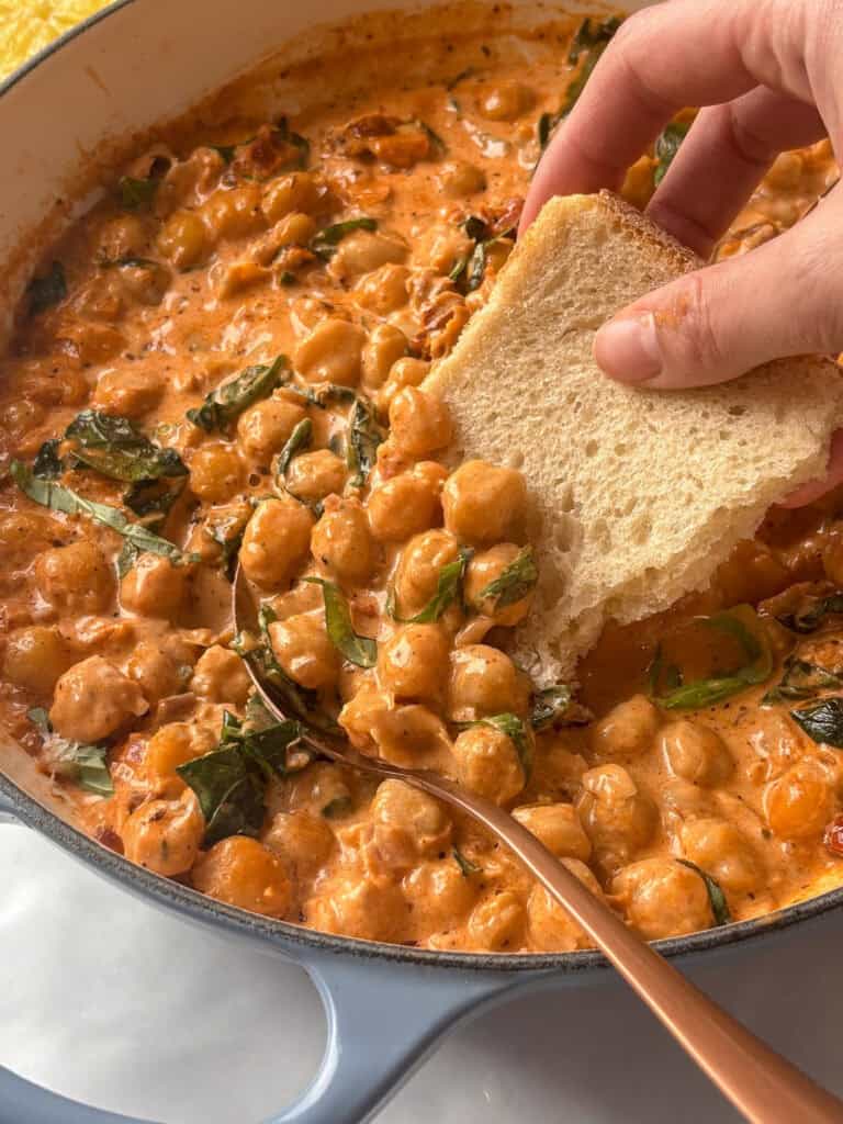 a casserole pan of marry me chickpeas, with sun-dried tomatoes, spinach, and basil in a creamy sauce. A slice of sourdough is taking a portion of the creamy chickpeas out the pan