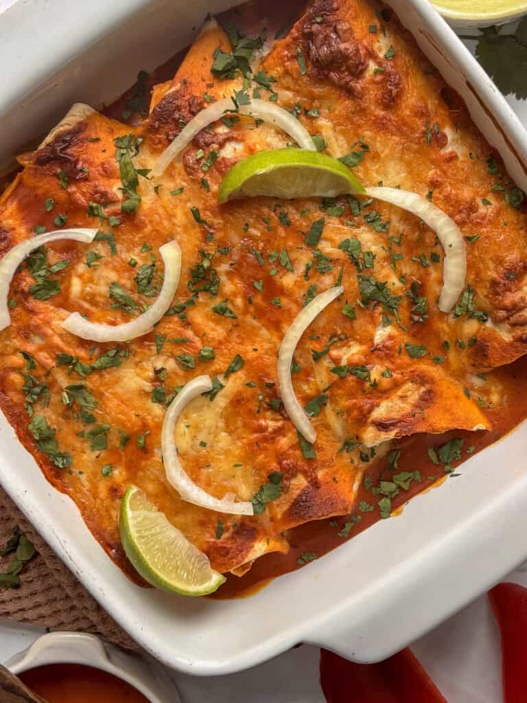 a white dish of black bean enchiladas topped by fresh cilantro, lime wedfes, and sliced onion