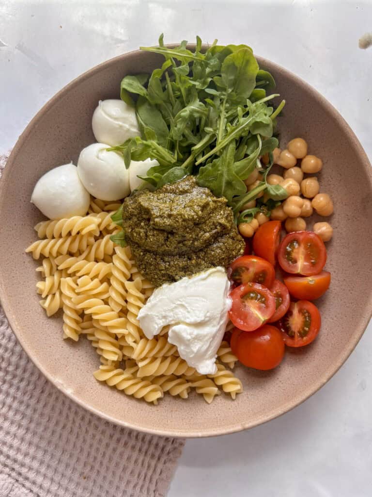 a bowl with the ingredients for pesto pasta with the cooked pasta, mozzarella pearls, arugula, chickpeas, cherry tomatoes, pesto and Greek yoghurt