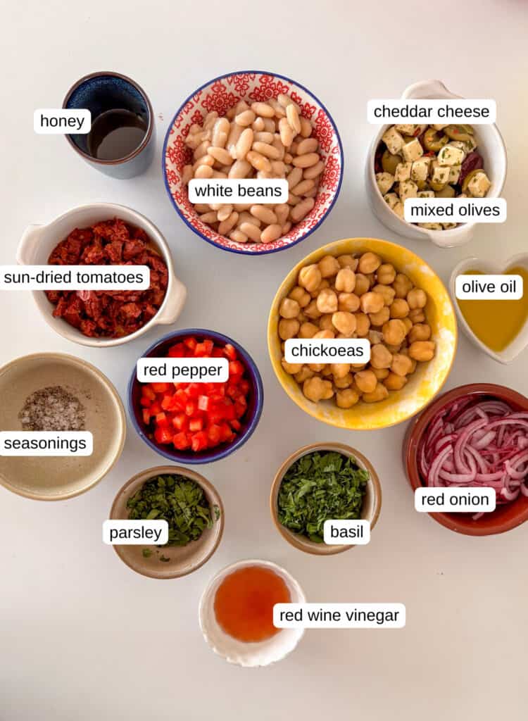 the full ingredients needed for this dense bean salad recipe, in separate bowls. Includes white beans, mixed olives, chickpeas, olive oil, red onion, red pepper, sun-dried tomatoes, parsley, red wine vinegar, seasonings