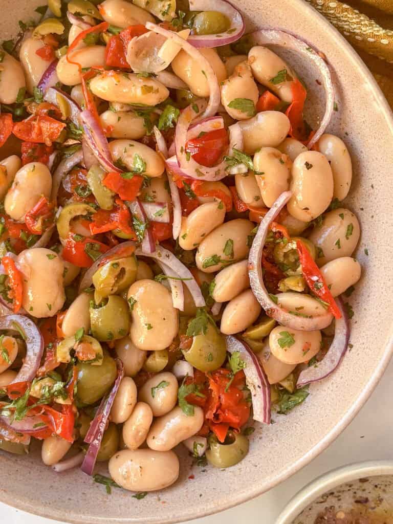 a white serving plate of marinated white beans with red peppers, onion, olives, and parsley