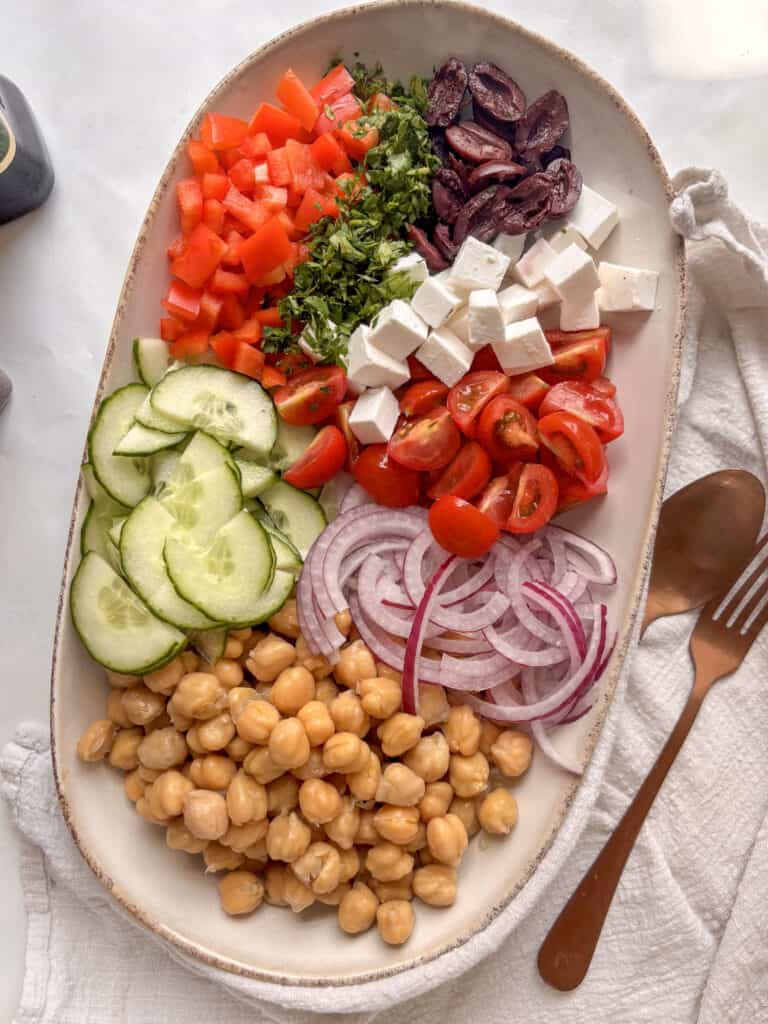 an oval serving platter with the ingredients for the mediterranean chickpea salad arranged separately on the plate on a white table