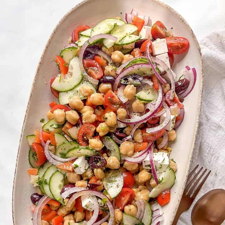 a serving plate of mediterranean chickpea salad with chickpeas, red pepper, tomatoes, red onion, olives, cucumber, and more