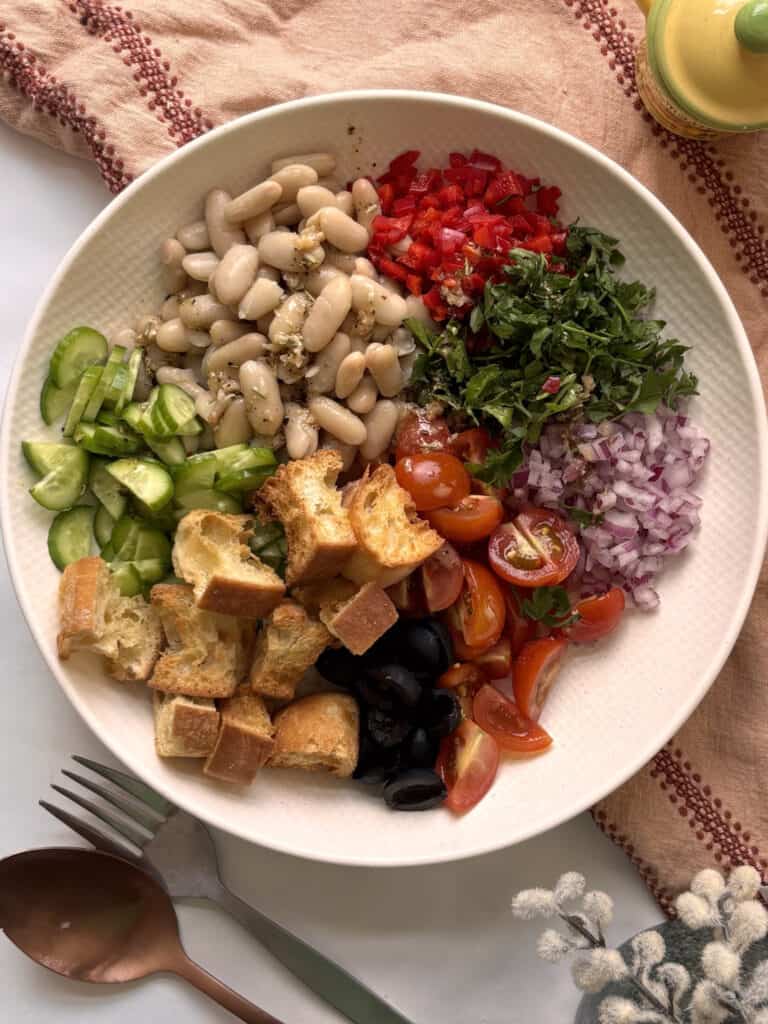 the ingredients needed for Italian white bean salad with the ingredients placed separately in the bowl - includes white beans, peppers, parsley, onion, tomato, olives, croutons, cucumber