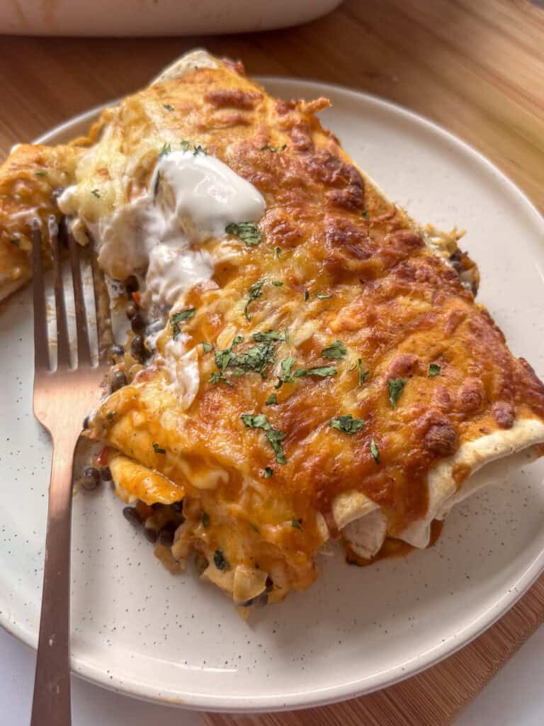 a close up of a serving of two lentil enchiladas on a white plate, topped with a little sour cream. A fork rests on the plate with a bite cut out of one encihlada