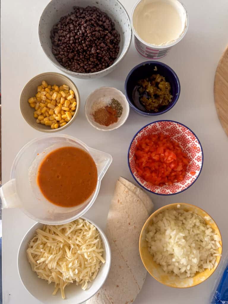 all of the ingredients needed to make lentil enchiladas: black lentils, sour cream, jalapenos, sweetcorn, spices, enchilada sauce, red peppers, onion, tortilla wraps, cheese