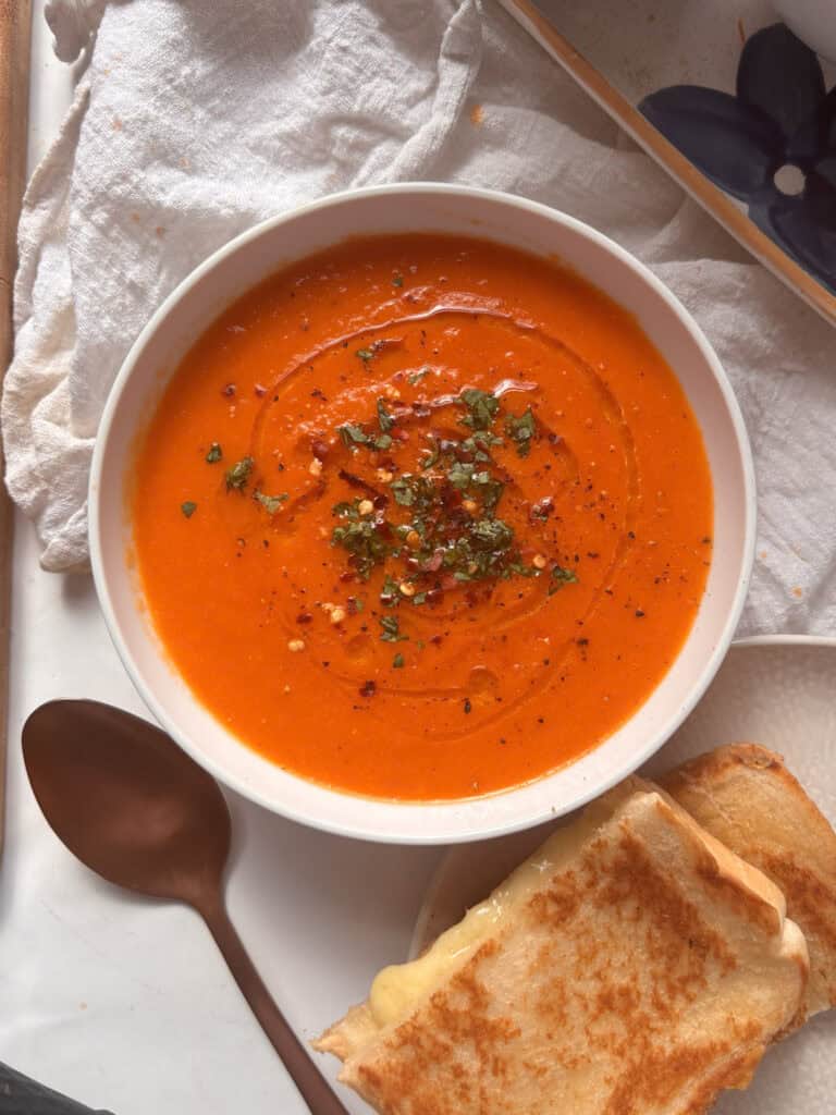 a white bowl of roasted red pepper and tomato soup topped with fresh herbs, olive oil, and red pepper flakes. A cheese toastie sits to one side just out of shot.