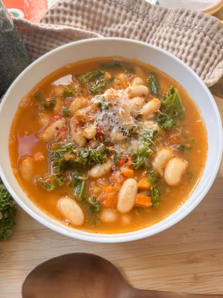 a white bowl of Tuscan white bean soup topped with Pecorino cheese, along with kale and white beans