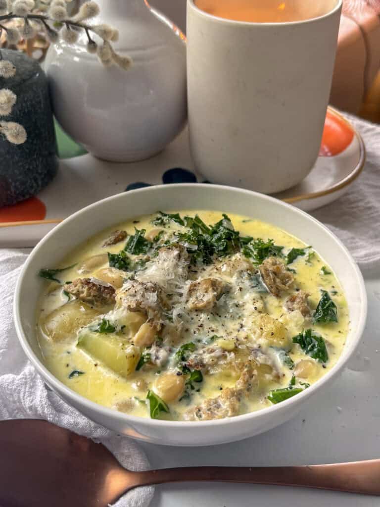 zuppa toscana - tuscan creamy sausage and bean soup with potatoes and kale, in a white bowl topped with hard cheese and black pepper