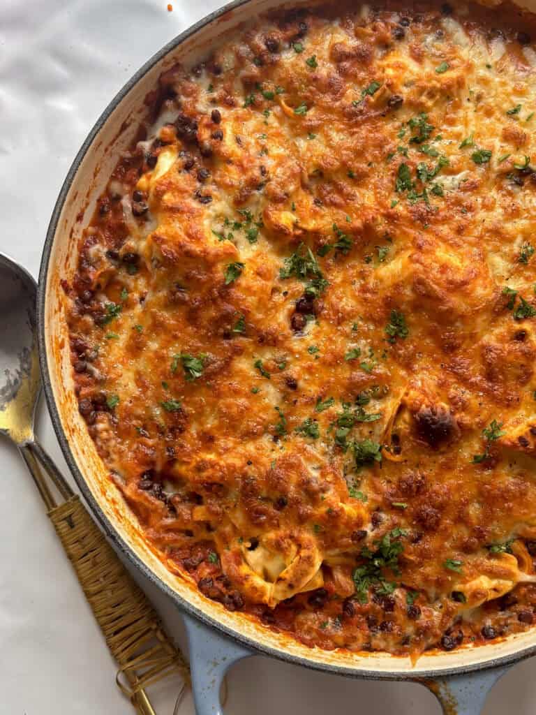 a close up shot of the pan of baked tortellini lasagna, showing the lentil ragu, tortellini, and a golden cheesy topping