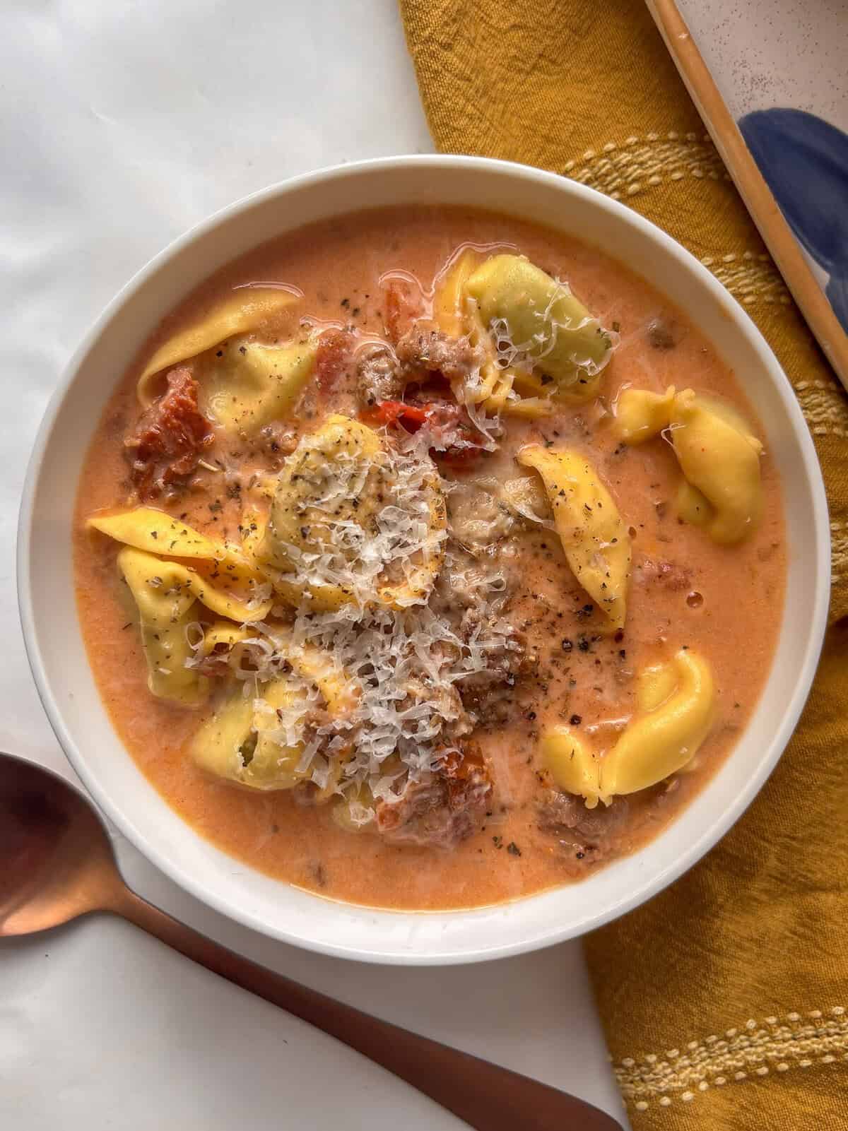 a white bowl of cozy marry me tortellini soup topped with black pepper and pecorino cheese