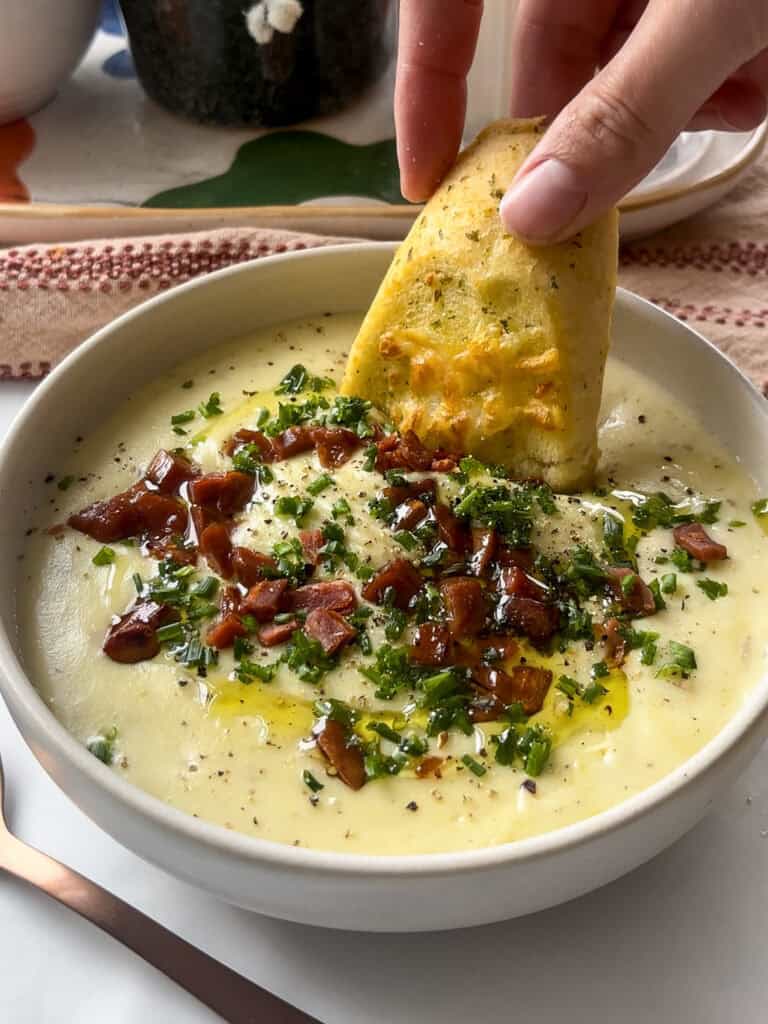 a bowl of potato and leek soup topped with vegan bacon and chives, with a slice of garlic bread being dipped into the soup