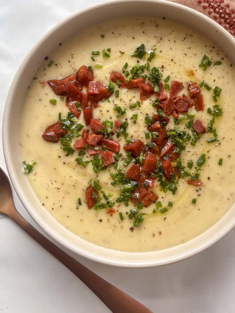 a close up photo of creamy potato and leek soup topped with crispy vegan bacon bits and chives, as well as black pepper and olive oil