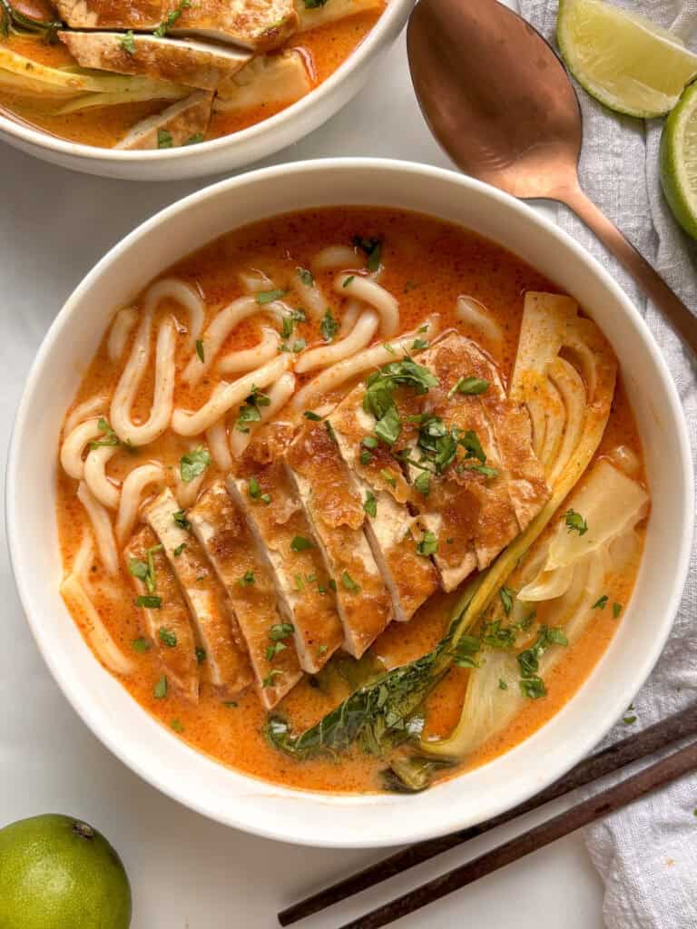a white bowl of red curry noodle soup topped with crispy sliced tofu and cilantro. Bok choi is also in the broth