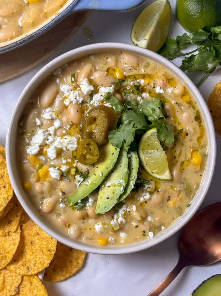a white bowl of white bean chili topped with feta, avocado, a lime wedge, and fresh cilantro. A scattering of tortilla chips can be seen on the table around the bowl of chili