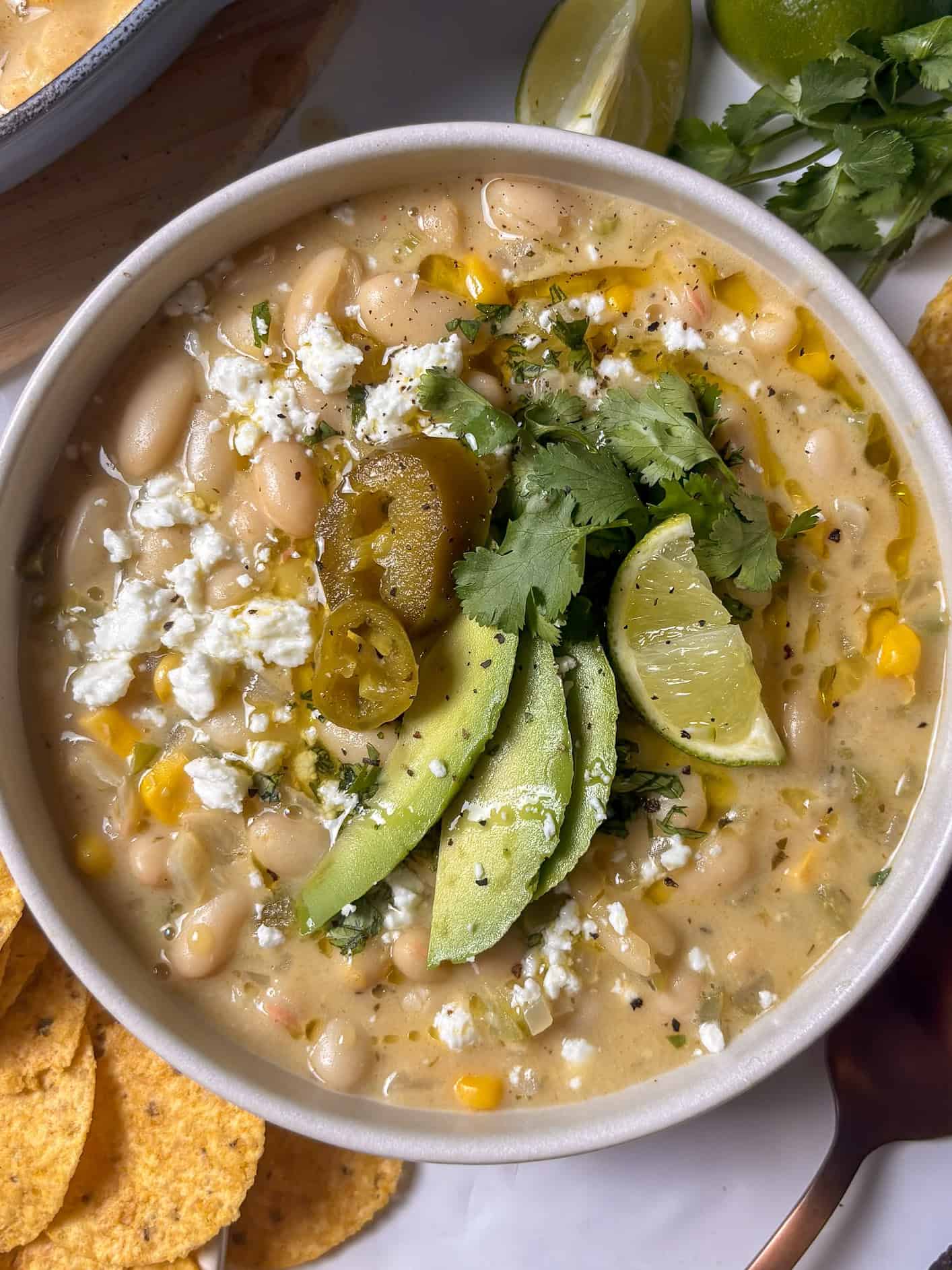 a close up shot of a creamy white bean chili loaded with sliced avocado, jalapenos, coriander, and lime