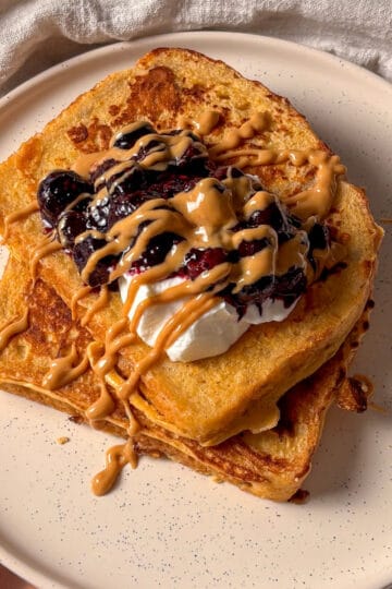 protein French toast stacked on a white plate, topped with greek yogurt, stewed blueberries, and a drizzle of peanut butter