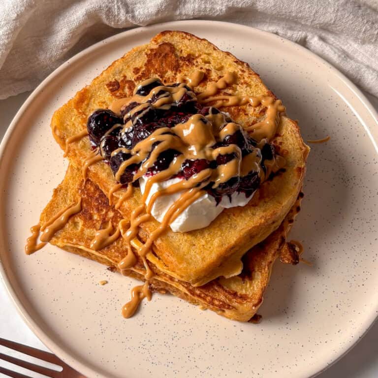 protein French toast stacked on a white plate, topped with greek yogurt, stewed blueberries, and a drizzle of peanut butter