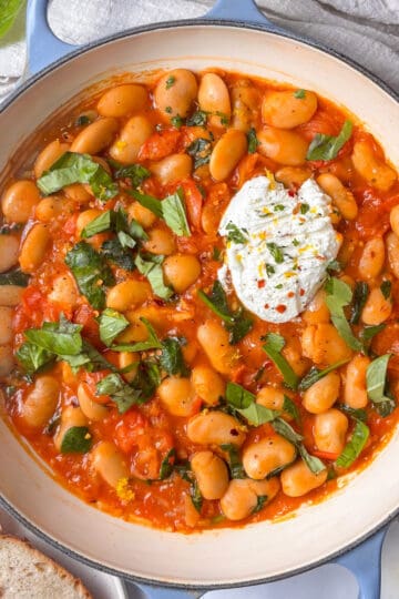 a casserole pan of cherry tomato ricotta butter beans, with a scoop of ricotta, fresh basil, and topped with lemon zest and red chili flakes