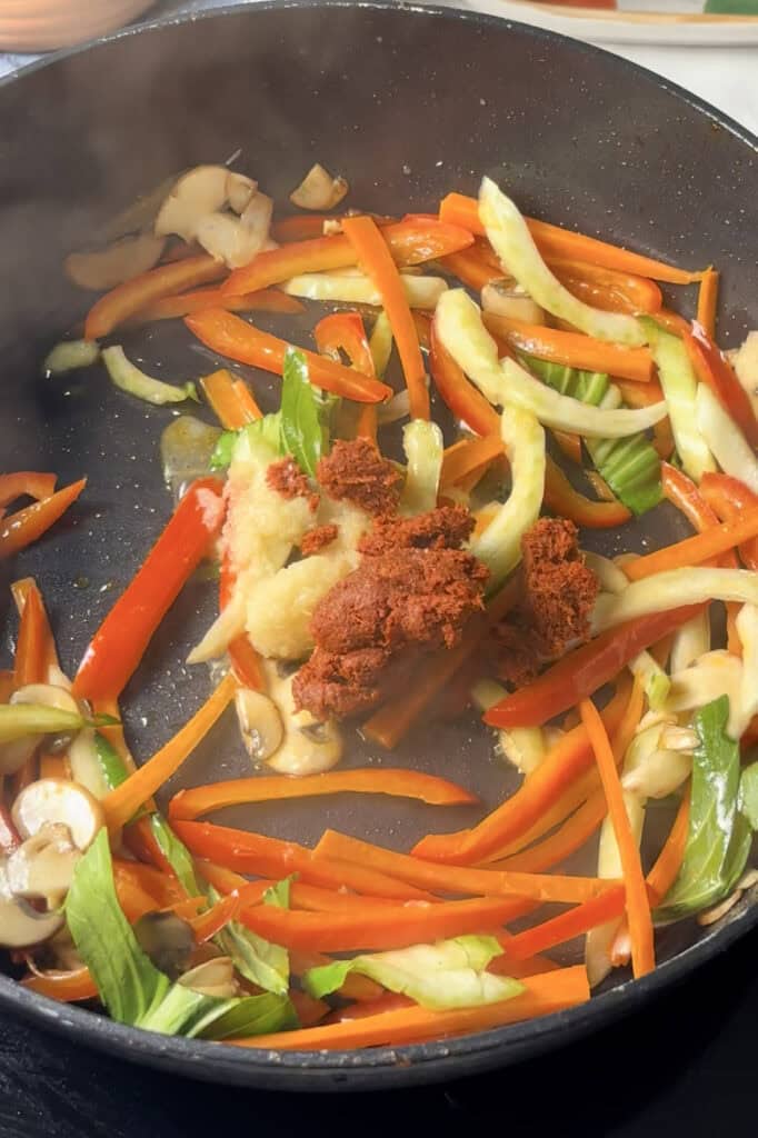 minced garlic, ginger, and red curry paste is added to a sauce pan of stir fried vegetables including peppers, mushrooms, and bok choi