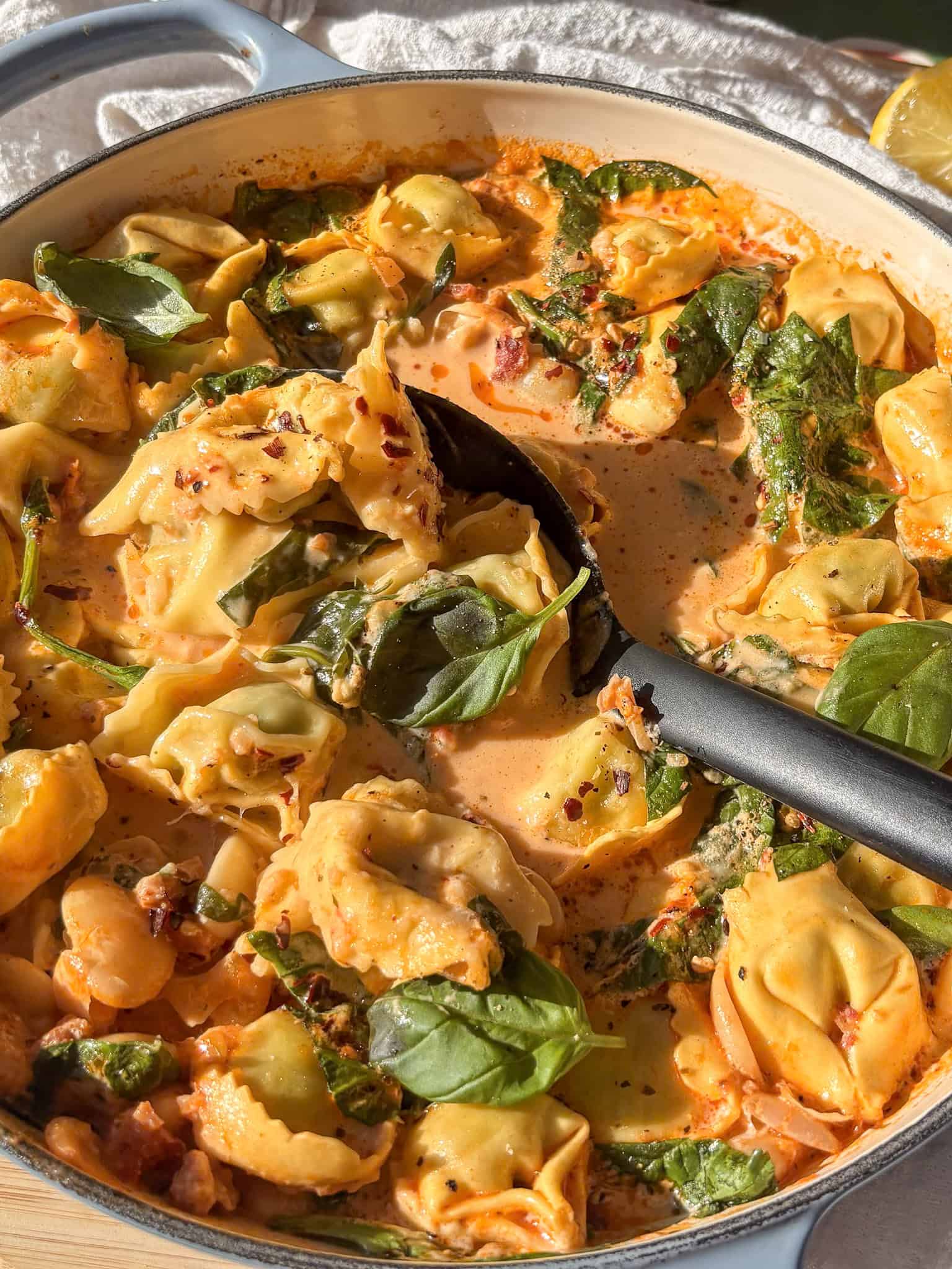 a pan of Marry Me tortellini with a spoon taking a scoop out of it. It's cast in direct sunlight, and topped with red chili flakes and fresh basil leaves