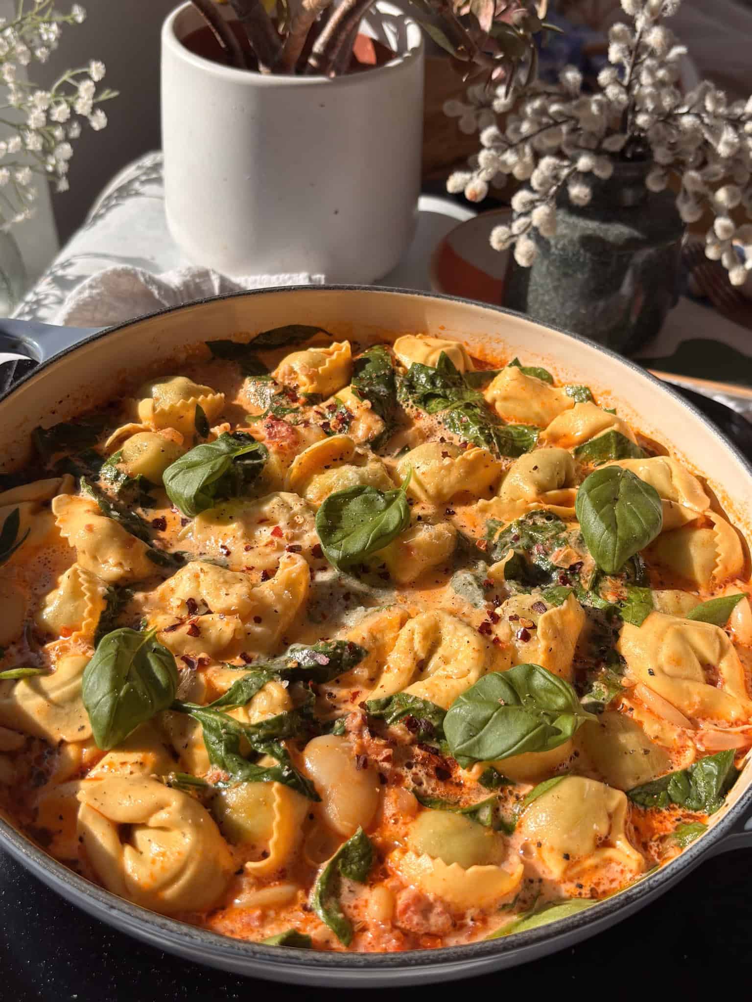a photo of a finished dish of Marry Me tortellini, with cooked tortellini and butter beans nestled in a creamy spinach and sun-dried tomato sauce, topped with red chili flakes and fresh basil.
