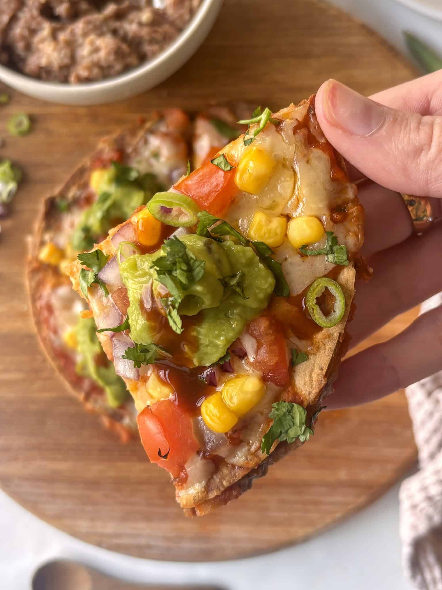 a quarter of a Mexican pizza, with in the background Mexican pizza on a chopping board, topped with diced tomatoes, sweetcorn, spring onion, red onion, and a spoonful of guacamole with diced cilantro and hot sauce