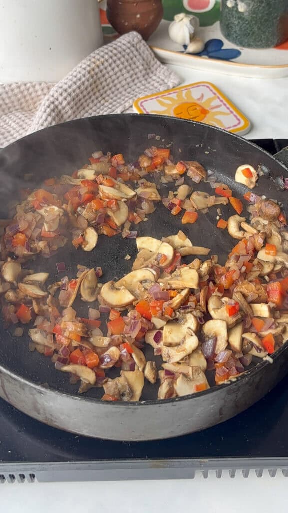 diced red onion, red pepper, garlic, and sliced mushrooms being cooked in a saucepan until softened
