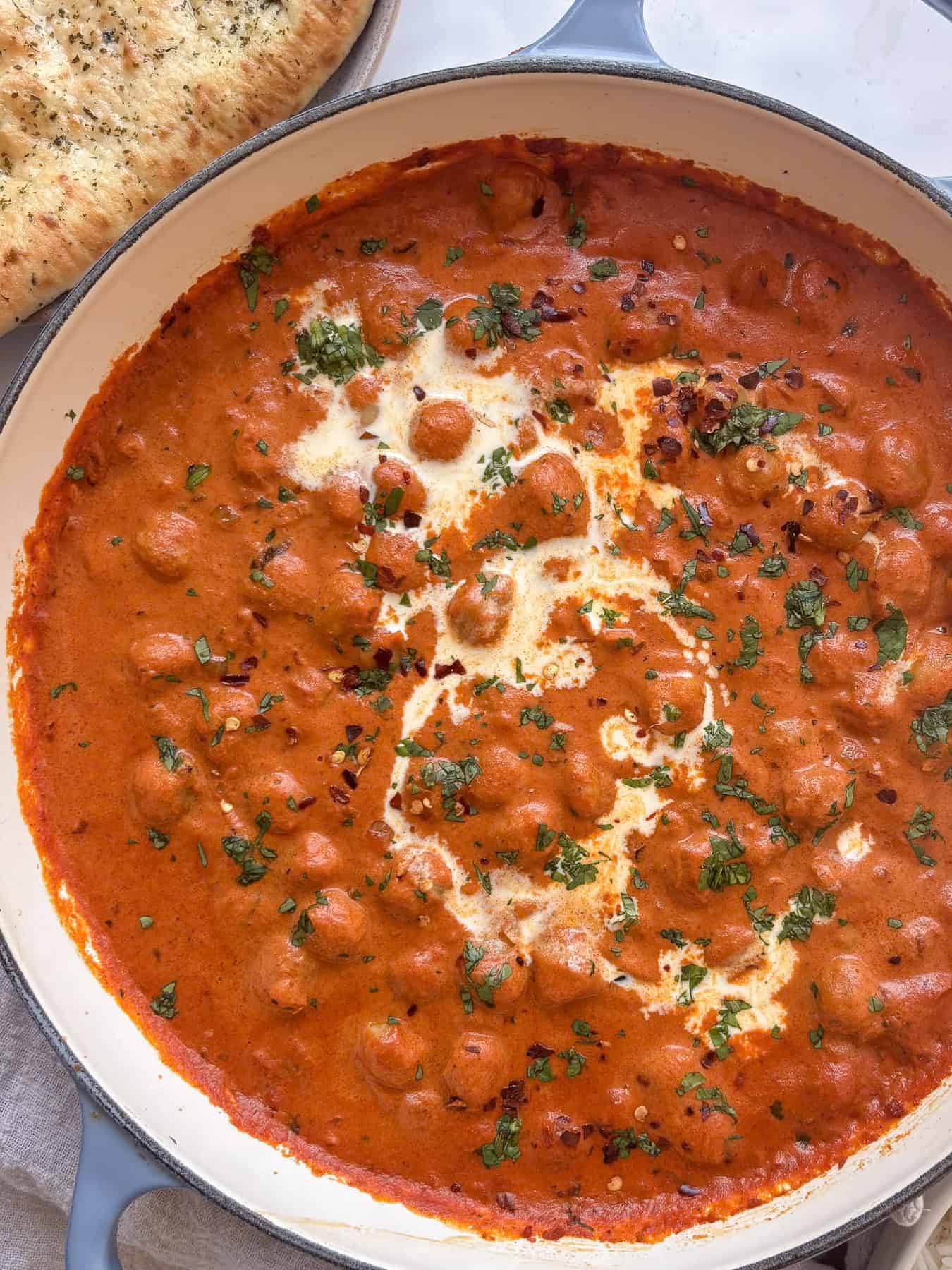 creamy chickpea tikka masala topped with cream, cilantro, and red chili flakes. A naan sits to one side of the pan.